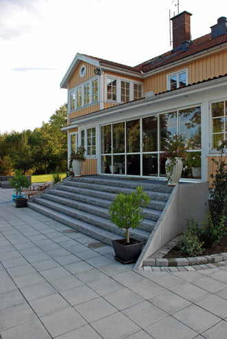 Stor och maffig trappa i svensk Bohuslänsgranit med råkilade framkanter och flammade ovansidor framför ett hus. Skapar en imponerande entré. Stengrossen säljer produkten.