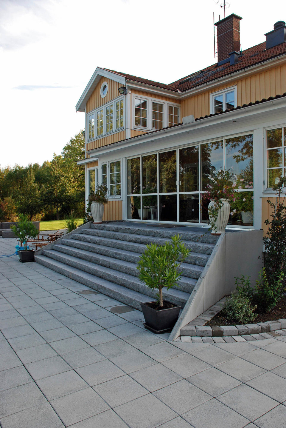 Stor och maffig trappa i svensk Bohuslänsgranit med råkilade framkanter och flammade ovansidor framför ett hus. Skapar en imponerande entré. Stengrossen säljer produkten.
