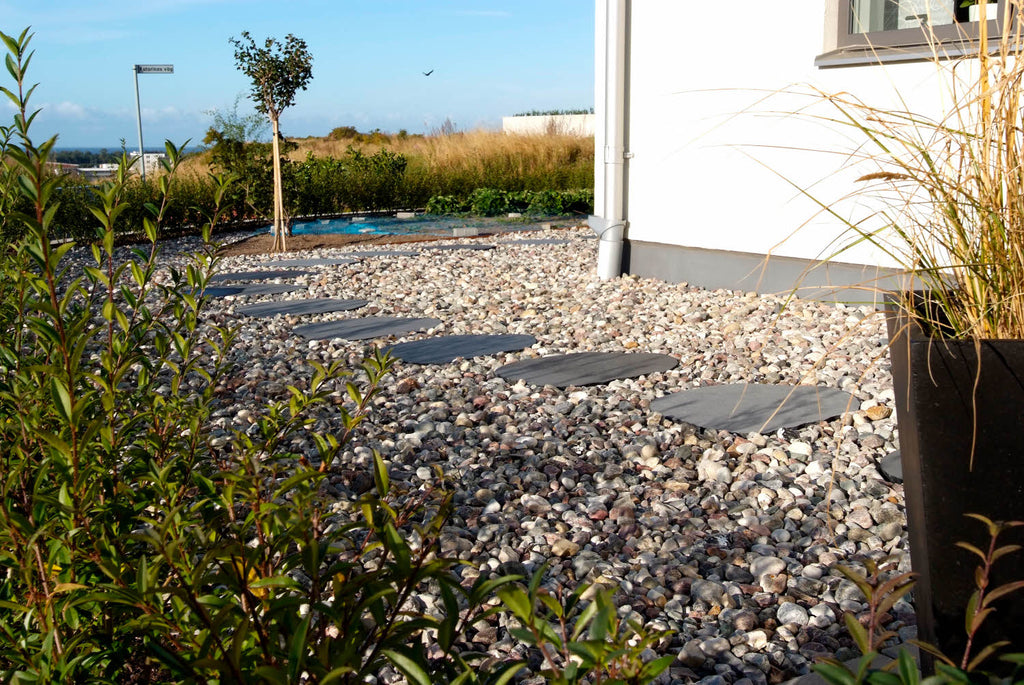 Dekorsten. Gul Sjösten. Det ligger trampstenar i skiffer bland dekorstenen. Ett hus finns i bakgrunden. Göna växter finns både i framkant som bakkant av bilden. Säljs av Stengrossen.se