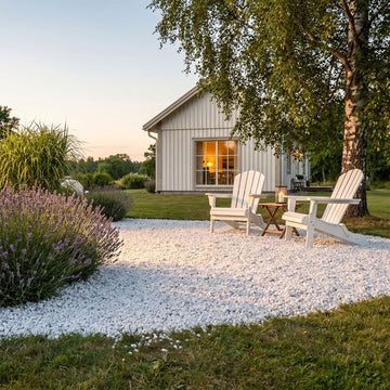 En tumlad dekorssten vid namn Bianco Carrara ligger mitt i en gräsmatta framför ett fritidshus i Sverige. Bland stenarna står ett par vita fritidsstolar med ett litet bord mellan sig. I bakgrunden syns en björk och lite blommor vid sidan. Stengrossen säljer produkten.