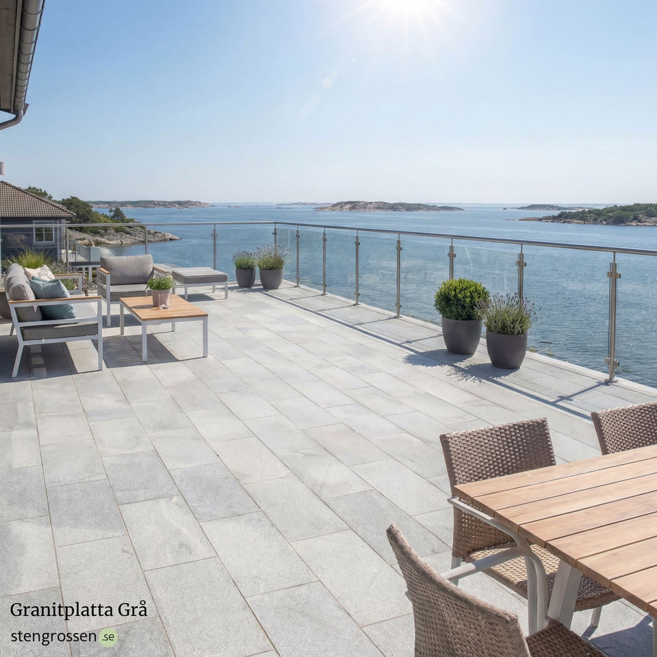 Granitplatta i grått på altan vid skärgården med soligt ljus, moderna fritidsmöbler och blommor samt glasräcke runt uteplatsen; Stengrossen säljer produkten.