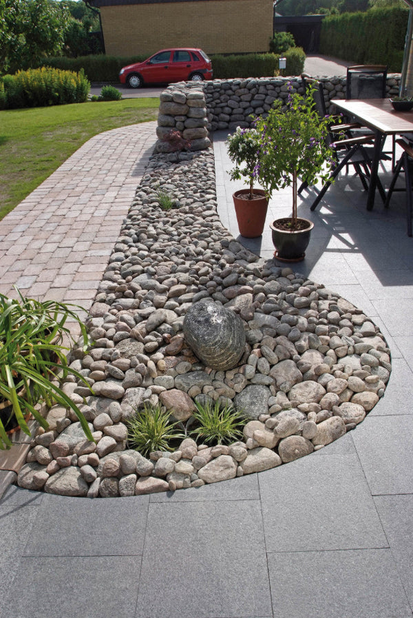 En trädgård med granitplattor, kullersten och marksten i betong. Ovanpå plattorna i granit står det ett bord, sstolar och ett par krukor. I bakgrunden är det en stenmur med kullersten. Säljs av Stengrossen.se