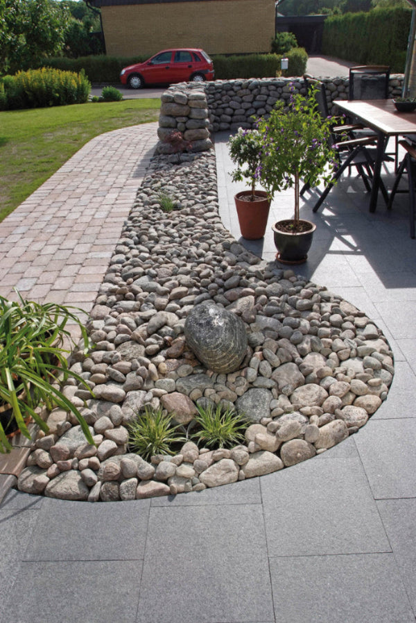 På mitten av bilden ligger det kullersten med ett par gröna växter. I nederkan, samt till hger i bilden ligger det stenplattor av granit. Till vänster i bild ligger det marksten av betong. Säljs av Stengrossen.se