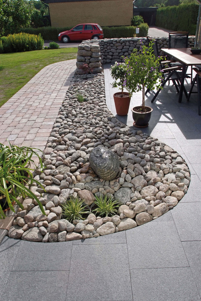 Stenplattor i granit. På insidan av plattorna ligger det kullersten. Bland kullerstenen finns det ett par gröna växter. Färgen på granitplattorna är grafit. Säljs av Stengrossen.se