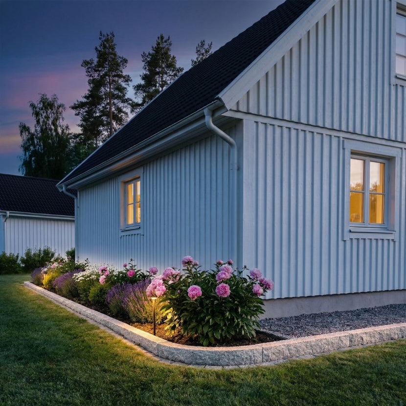 Granitkantsten som kallas för GV6 ramar in en blomsterbädd framför ett hus. På andra sidan av kantstenen är det en gräsmatta. GV6 har en storlek av 100x20x8 cm och är grå. Bilden är tagen i skymningsljus. Stengrossen säljer kantsten.