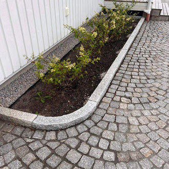 Kantsten i granit. GV6. Ligger längs med ett vitt hus. Mellan huset och kantstenen är det en blomsterbädd. På andra sidan ligger det smågatsten. Stengrossen säljer kantsten och smågatsten.