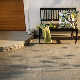 Grå granitplatta tillverkad i Portugal framför ett vitputsat hus med en svart bänk och kuddar ovanpå plattorna, samt en träfärgad ytterdörr. Stengrossen säljer produkten.