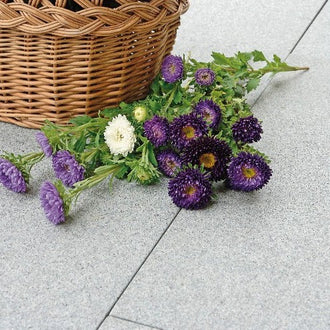 Grå granitplatta från Portugal med en korg fylld med lila, vita och gröna blommor ovanpå, samt snittblommor bredvid korgen. Kontrasten mellan den grå stenen och de färgglada blommorna syns tydligt. Stengrossen säljer produkten.