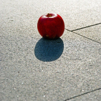 Närbild av grå granitplatta från Portugal med synlig kornstorlek på den flammade ytan, och ett rött äpple placerat ovanpå plattan i bildens övre del. Stengrossen säljer produkten.