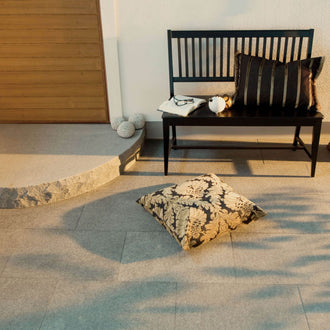 Granitplatta Porto grå framför ett vitputsat hus, med en svart bänk och kuddar ovanpå samt en beige och svart kudde direkt på plattan. Stengrossen säljer produkten.