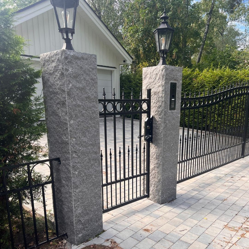 Två grå granitstolpar i bohusgranit står vid infarten till ett garage. Mellan stolparna finns en smidesgrind för gångtrafik, och till höger om den ena stolpen finns en bred smidesgrind för bilinfart. Lykttor är monterade ovanpå varje stolpe. Stengrossen säljer produkterna.