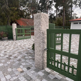 Två grå granitstolpar markerar infarten till ett hus med garage. På stolparna hänger gröna trägrindar. Storgatsten ligger på marken från vägen och fram mot garaget. Stengrossen säljer produkterna.