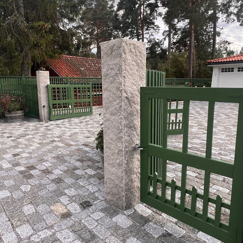 Två grå granitstolpar markerar infarten till ett hus med garage. På stolparna hänger gröna trägrindar. Storgatsten ligger på marken från vägen och fram mot garaget. Stengrossen säljer produkterna.