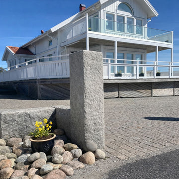 En grå granitstolpe i Bohusgranit med måtten 130x30x30 cm markerar infarten till ett modernt vitt hus. Vid infarten finns grå bohus smågatsten, och en blomkruka med gula blommor står ovanpå stolpen. Stengrossen säljer produkten.
