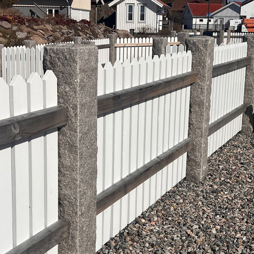 Ett antal grå granitstolpar i bohusgranit, 170x20x20 cm, med råkilad yta, totalt tio stycken. Mellan varje stolpe är ett vitt trästaket monterat. På marken nedanför stolparna ligger det dekorsten i form av makadam. Stengrossen säljer produkterna.