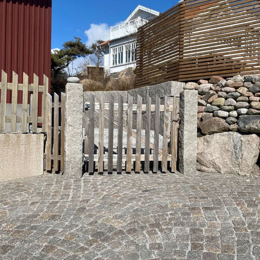 Två granitstolpar i grå bohusgranit markerar en infart för gångtrafik. Mellan stolparna finns en träfärgat trägrind. Till höger om stolparna syns en kullerstensmur, och till vänster ett träfärgat trästaket. I bakgrunden står en granitmur med staplae block i grå bohus. Stengrossen säljer produkterna.