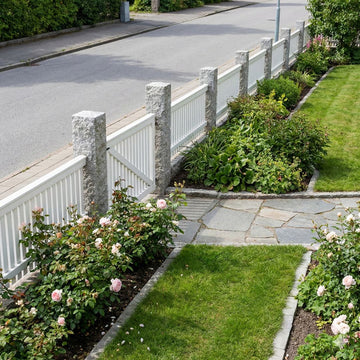 Granitstolpar i färgen grå som står längs med en väg. Mellan stolparna är det ett vitt staket. På insidan av stolparna och staketet är det en trädgård. Stengrossen säljer granitstolparna.