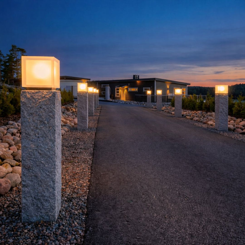 Infart mot hus med asfalterad väg, flankerad av grå granitstolpar i storleken 170x30x30 cm. Ovanpå stolparna sitter lampor som lyser upp i skymningen. Stengrossen säljer produkten.