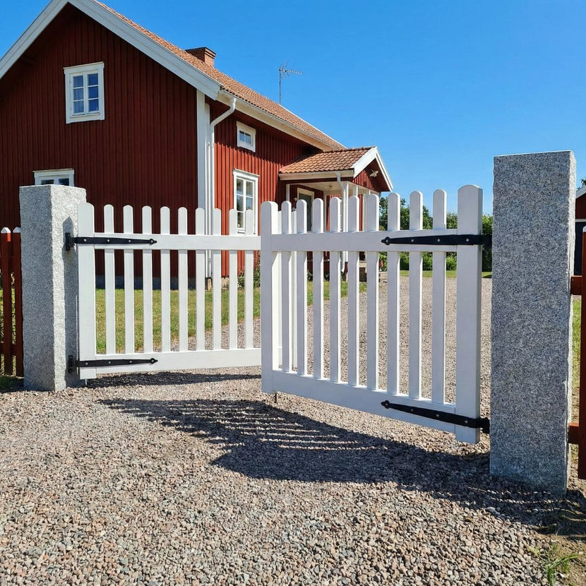 Grå granitstolpar i storleken 170x30x30 cm står som portstolpar vid infarten till ett rött hus. Mellan dessa stolpar finns en vit tvådelad grind med svarta gångjärn. Infarten är grusad. Stengrossen säljer produkten.