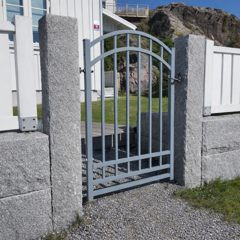 Svensktillverkade granitstolpar i grå bohus, 170x20x20 cm, med en grå grind mellan dem. Intill finns en grå granitmur och ovanpå muren står ett vitt staket. Stengrossen säljer produkten.