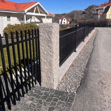 Granitstolpe i grå Bohusgranit. Måtten är 170x30x30 cm. Ytan är råkilad. På ena sidan av granitstolpen är det monterat ett staket i svart smide. På andra sidan är det en svart grind i svart smide. På insidan är det ett vitt hus. Stengrossen säljer produkten.