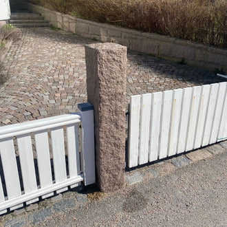 En röd granitstolpe i bohusgranit, 170x25x25 cm, har en råkilad yta. På båda sidor om stolpen är ett vitt trästaket monterat. Ovanför stolpen ligger smågatsten i olika kulörer, och längre upp ovanför smågatstenen finns en granitmur i samma röd bohusgranit. Stengrossen säljer produkterna.