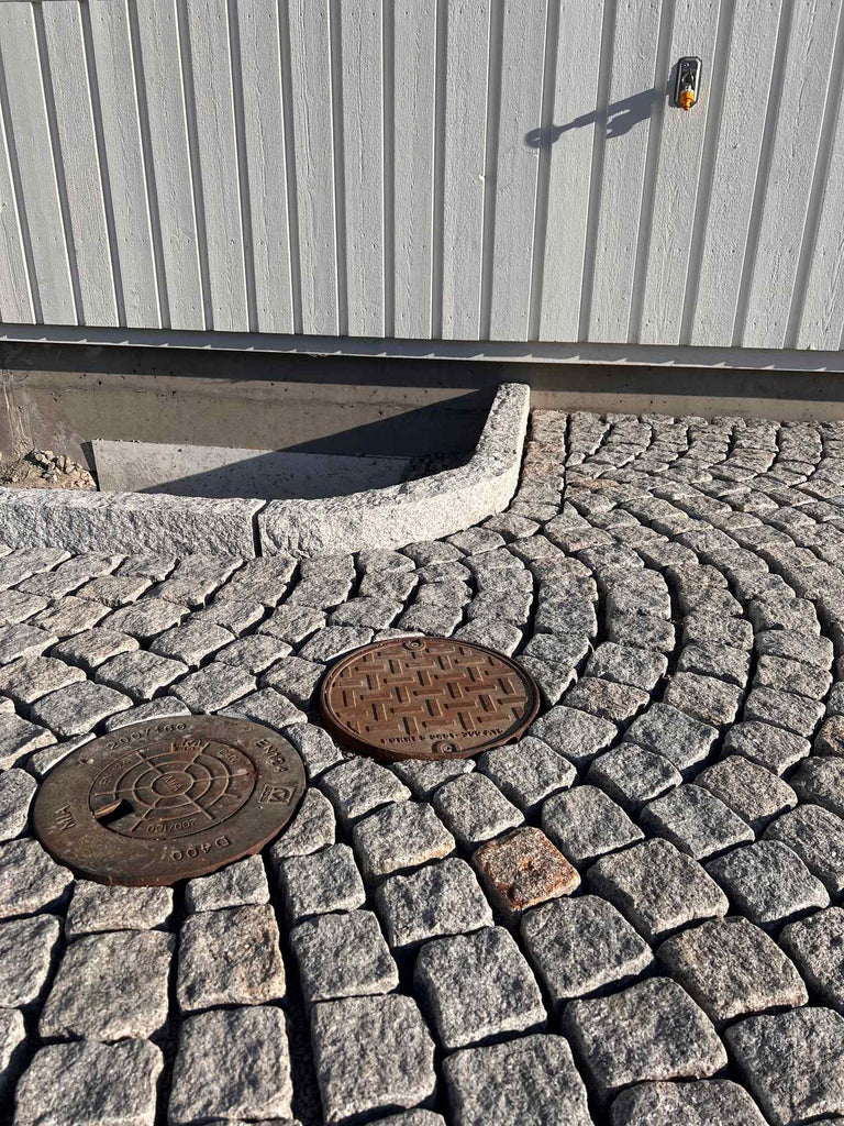 Gatsten som är lagda runt två stycken brunnslock. I bakgrunden är det ett hus. Gatstenen hålls på plats av kantsten i granit. Kantstenen kallas för GV4. Säljs av Stengrossen.se