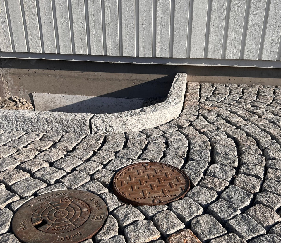Gatsten (smågatsten) som är av sorten Grå Bohus. Kantsten av sorten GV4 håller gatstenen på plats. I bakgrunden är det en husvägg med vit träfasad. I nederkant är det två brunnslock. Säljs av Stengrossen.se