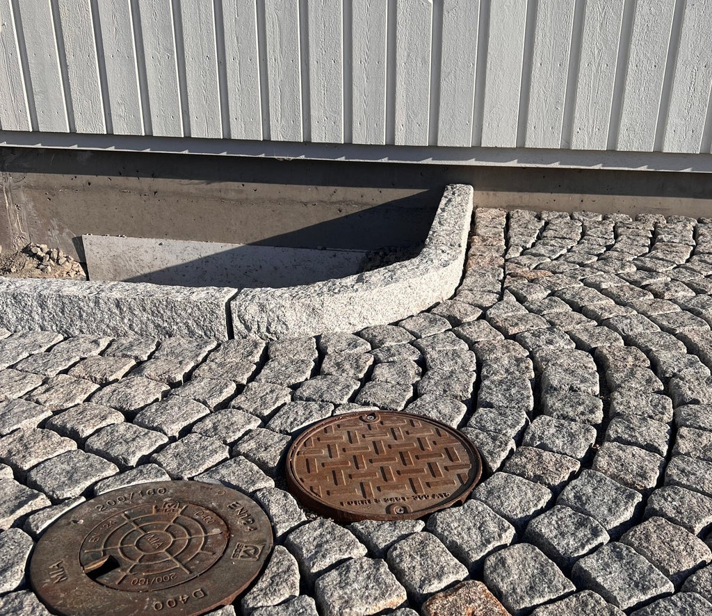 Gatsten (smågatsten) som är av sorten Grå Bohus. Kantsten av sorten GV4 håller gatstenen på plats. I bakgrunden är det en husvägg med vit träfasad. I nederkant är det två brunnslock. Säljs av Stengrossen.se