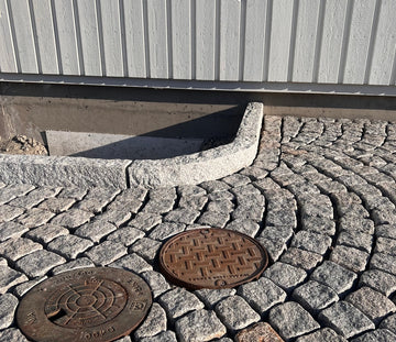 Gatsten (smågatsten) som är av sorten Grå Bohus. Kantsten av sorten GV4 håller gatstenen på plats. I bakgrunden är det en husvägg med vit träfasad. I nederkant är det två brunnslock. Säljs av Stengrossen.se