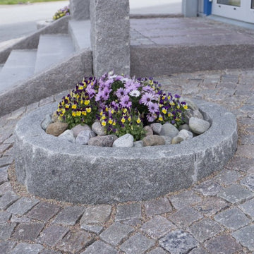 Den här bilden visar en radiekantsten i sorten grå bohus som formar en cirkel. Inuti cirkeln finns det dekorstenar, och på utsidan av cirkeln syns smågatsten. Stengrossen säljer produkten.
