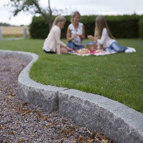 Kantsten i granit, sorten Grå Bohus som ramar in en gång med grus. På ovansidan av kantstenen är det en gräsmatta med personer som fikar på en filt. Stengrossen säljer kantstenen.