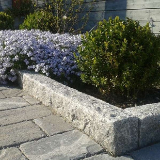 Granitkantsten i färgen Grå Bohus som ramar in en blomplantering. På utsidan ligger det grå marksten i betong. Stengrossen säljer produkterna.
