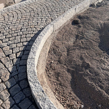 Kantsten GV4 i grå granit. På ena sidan av kantstenen ligger det smågatsten och på andra sidan kan man se betong som håller kantstenen på plats. Stengrossen säljer produkten.