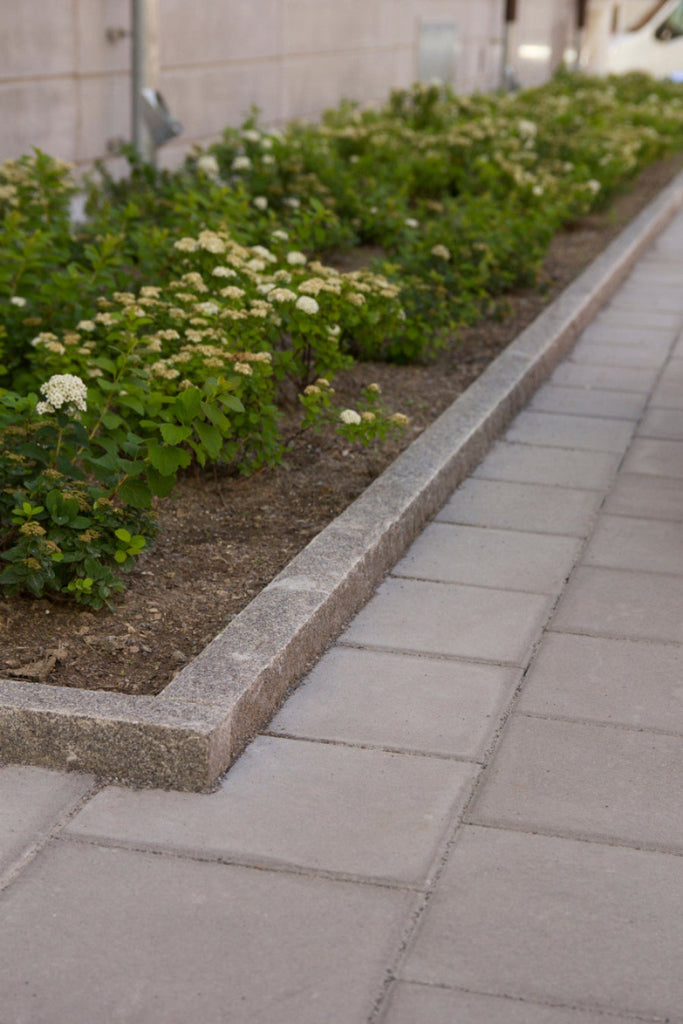 Granitkantsten RV4 i Grå Bohus, svensktillverkad och elegant. På utsidan ligger släta betongplattor, medan insidan ramar in en vacker blomplantering. Stengrossen säljer produkten.
