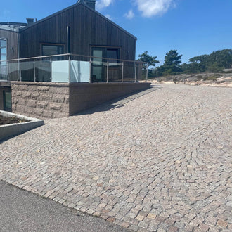 Infart med smågatsten Röd Bohus intill mur av röd beklädnadsgranit och hus med grå cederträfasad bakom – Stengrossen säljer produkten.