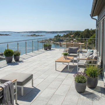 Solig uteplats vid havet belagd med grå granitplattor 60x30x3 cm med krukor med blommor och glasräcke i bakgrunden, Stengrossen säljer produkten.