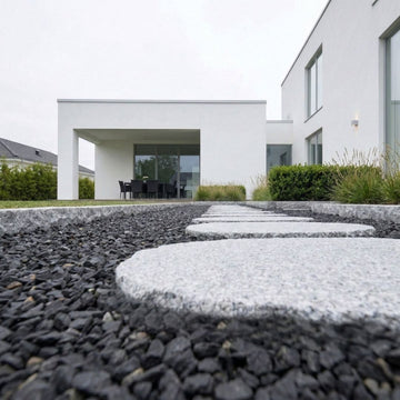 Steppingstones i granit, även kallade stegstenar eller trappstenar, lagda på rad i en gång intill ett vitt putsat hus och placerade i en bädd av svart diabas som skapar kontrast i trädgårdsmiljön. Stengrossen säljer produkten.