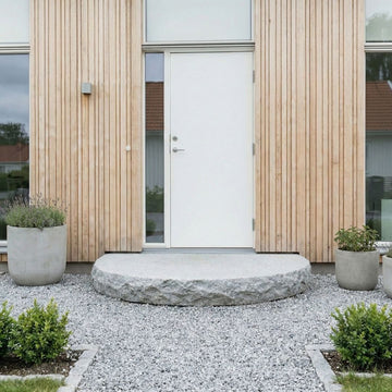 Entréplan i grå granit framför ett hus med träfärgad fasad, intill en vit dörr med glasparti till vänster, omgiven av gårdsgrus och krukor med växter. Stengrossen säljer produkten.