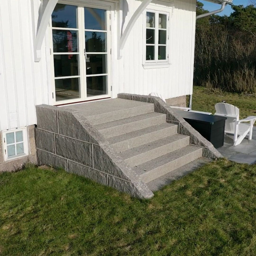 Granittrappa i Grå Bohus med fem steg som leder upp till dubbeldörrar på ett gammalt vitt trähus. Trappan ramas in av blockstensmurar i Grå Bohus på båda sidor. Produkterna säljs av Stengrossen.