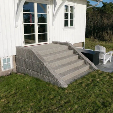 Granittrappa i Grå Bohus med fem steg som leder upp till dubbeldörrar på ett gammalt vitt trähus. Trappan ramas in av blockstensmurar i Grå Bohus på båda sidor. Produkterna säljs av Stengrossen.