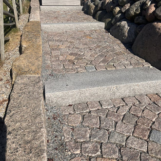 Svensktillverkade trappsteg i Grå Bohus, med smågatsten mellan trappstegen. Trappan ramas in av kantstenar på ena sidan och en mur av kullersten på den andra. Stengrossen säljer produkterna.