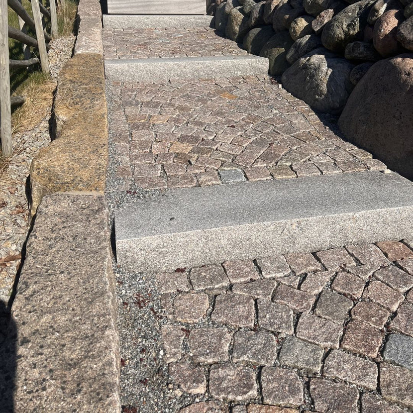 Svensktillverkade trappsteg i Grå Bohus, med smågatsten mellan trappstegen. Trappan ramas in av kantstenar på ena sidan och en mur av kullersten på den andra. Stengrossen säljer produkterna.