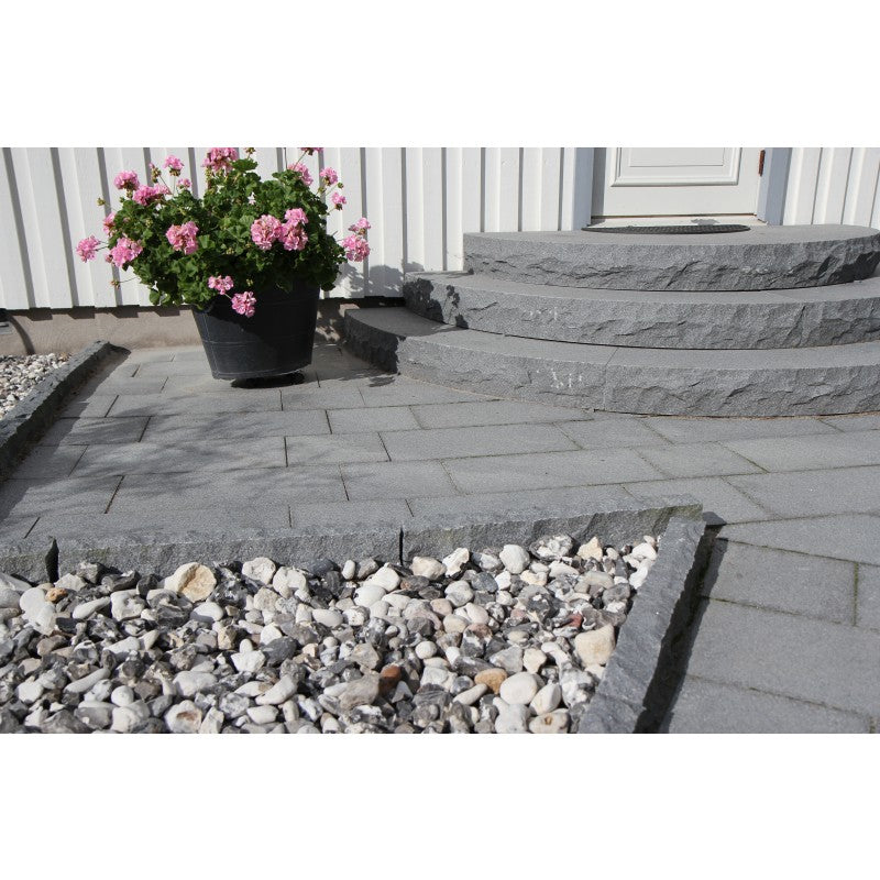 Trappa, kantsten och plattor i granit. Färgen är grafitgrå. Trappan har 3 steg. Plattorna är i två olika storlekar, 60x30x3 cm och 30x30x3 cm. På utsidan av kantstenen ligger det dekorsten som är av sorten Svartvit Sjösten. Ovanpå plattorna står det också en kruka med blommor. Säljs av Stengrossen.se