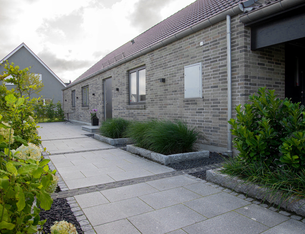 Svart gatsten tillsammans med granitplattor. Kantsten i granit ramar in rabatter med gröna växter. I bakgrunden är det ett hus med tegelfasad. Säljs av Stengrossen.se