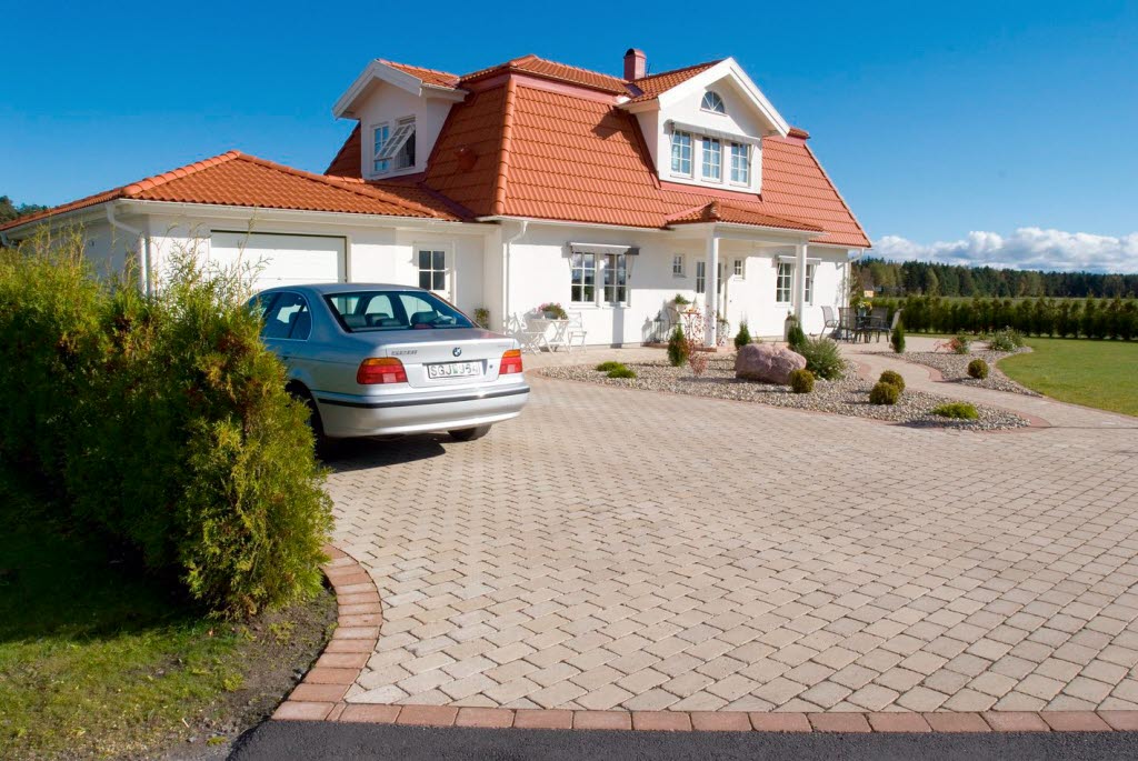 Karminröd marksten i serien Rustik från tillverkaren S:T Eriks. Den karminröda stenen fungerar som avgränsare mellan asfalten och den naturgråa markstenen.  Fint vitt hus med på bilden också.