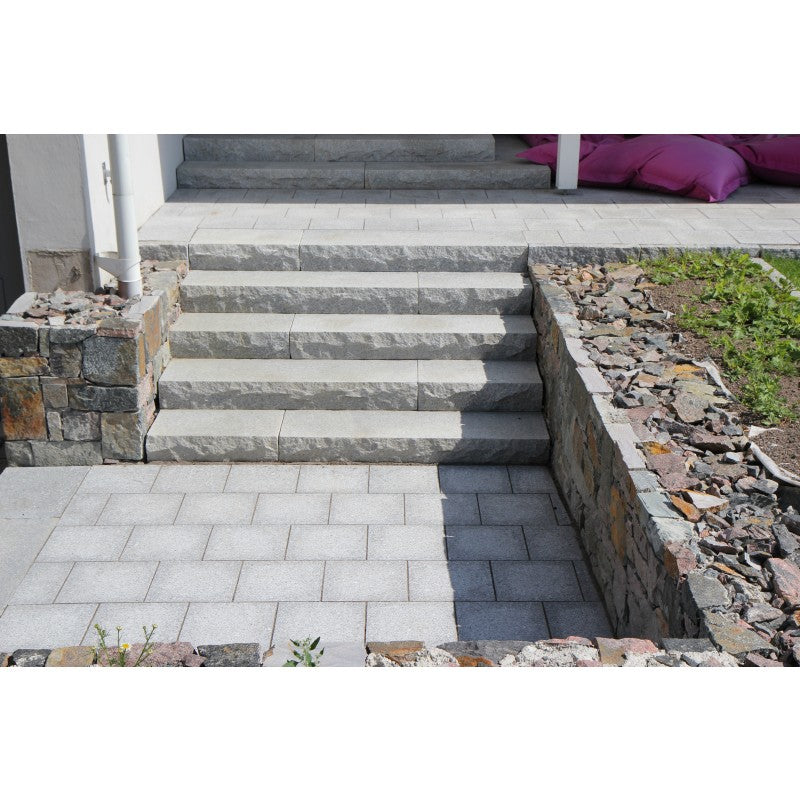 Grå granitplatta från Stengrossen. 30x30x3 cm. Plattorna ligger framför en granittrappa. Runt trappan och lattorna är det en sprängstensmur. Sprängstenen är i olika färger.